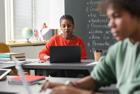 Portrait of Black young woman using laptop in school classroom with defocused students in foreground, copy space