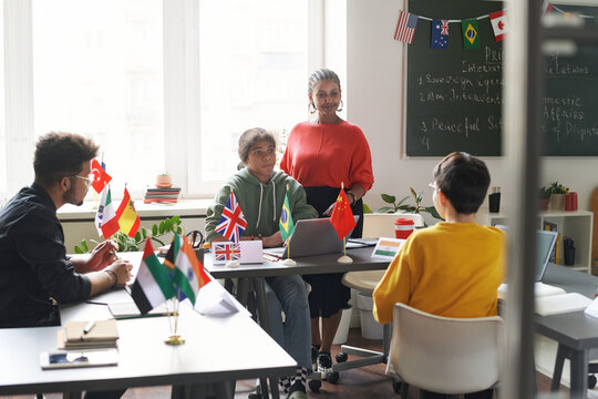 Diverse group of students sitting at table with flags during international conference in school classroom