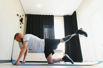 A man with a prosthetic leg does sports exercises on a mat at home. The concept of rehabilitation