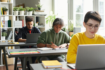 Portrait of young Black girl working in classroom while sitting at desk and taking notes, copy space