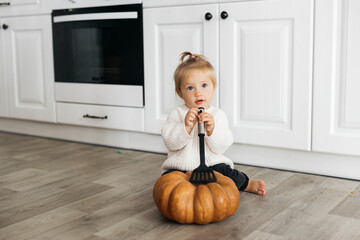 baby sits on the kitchen floor next to a large pumpkin and holds a kitchen spatula. He is wearing a...