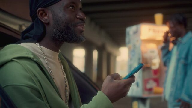 Black man leaning against open car trunk, checking social media on smartphone and smiling, his friend talking on retro payphone under urban overpass in background. Rack focus
