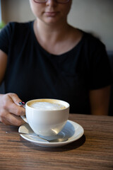 Front view of female hand holding white porcelain cup of cappuccino or latte coffee drink on brown wooden table in cafe or restaurant. Soft focus. Copy space. Hot drinks theme.