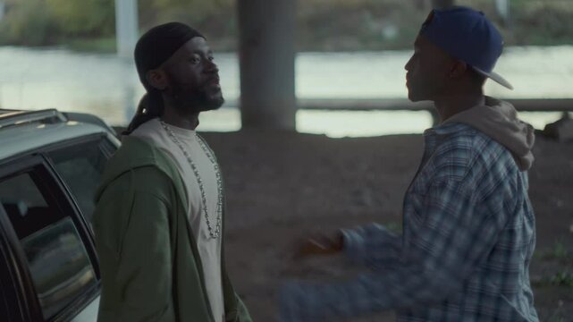 Two angry African American men having quarrel, gesturing vigorously, blaming and pushing each other near car parked under overpass with river in the background