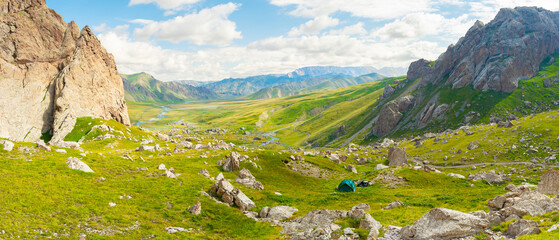 In the image, a green tent is set up at the foot of the cliffs under a blue sky with white clouds
