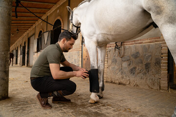 Young man preparing working bandages for his horse before training. Riding club concept