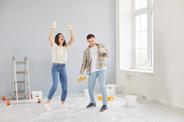 Fototapeta premium Happy young married couple have fun while doing apartment repair, painting wall with rollers. Cheerful spouses dancing together at their own new apartment, enjoying festive mood and celebrating.
