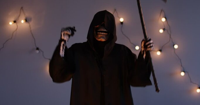 Close up shot of cheerful man in grim reaper death costume dancing with scythe in his hand. Man in long black cloak with hood and skull mask enjoying good mood, having fun and celebrating Halloween.