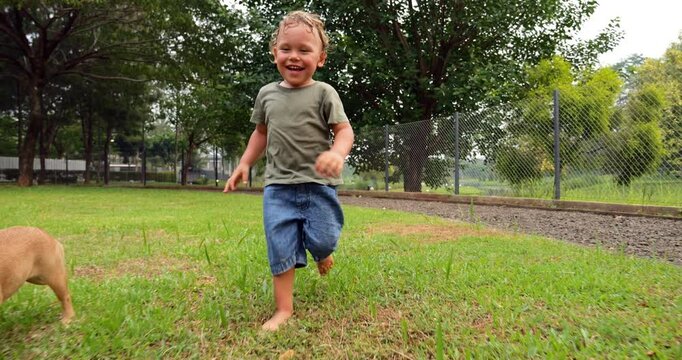 Barefoot and joyful toddler runs in fenced dog play area on hot summer day, accompanied by his French Bulldog puppy nearby. Slow motion shot, child's expressive face exudes happiness and excitement