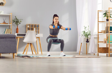 Woman engages in a home workout focused on fitness, performing squats and stretching exercises with a resistance band. Sport and training is evident as she maintains an effective exercise.