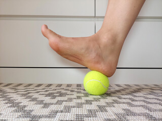 Woman is massaging her foot with a tennis ball on a rug