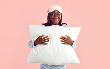 Happy young african american woman wearing sleep eye mask and sleepwear standing isolated on a studio pink background and hugging white soft pillow. Healthy sleep and good night concept.