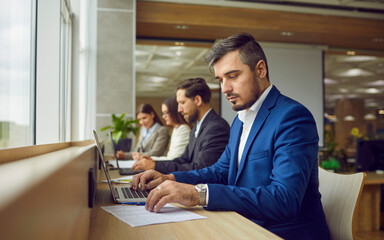 Serious business people sitting in a row and working on laptop in modern open plan office sitting at same desk. Company employees wearing suits typing on computers on their workplace.