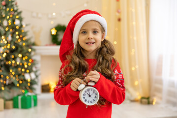 Little girl baby child holding white alarm clock. New Year's clock at midnight. Twelve O'clock midnight in retro style. Christmas decoration on wood background. a clock displays just before New Year