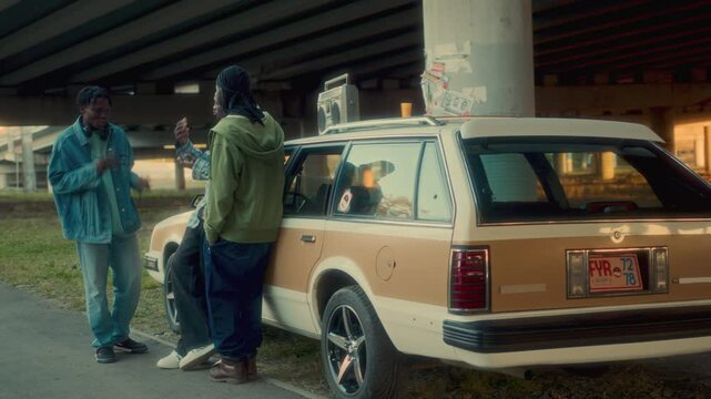 Group of cheerful African American friends in street style clothes having conversation beside retro station wagon with boombox on roof, parked under city bridge