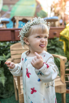 Crian&ccedil;a branca loira com enfeite de flores no cabelo, fazendo uma careta engra&ccedil;ada com vestido de estampa