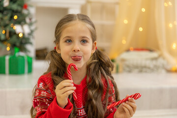 Happy little girl with a lollipop near Christmas tree at home. Little girl with sweet candies on Xmas eve. Family, tradition, celebration concept. Candy canes in shape of heart in hand child