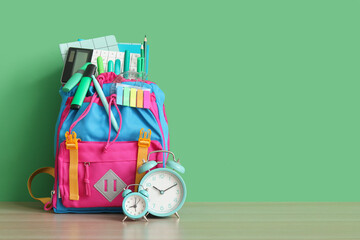 Colorful school backpack with different stationery and alarm clocks on wooden table near green wall