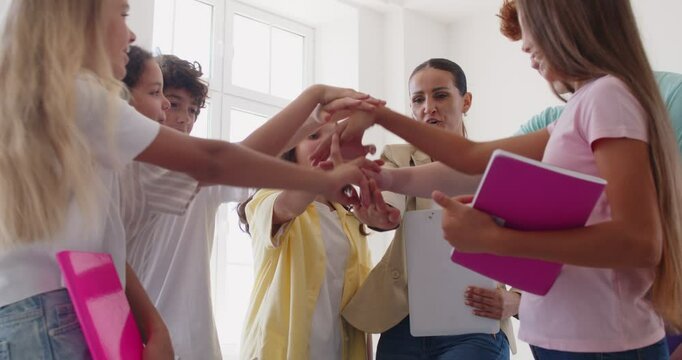 Students and female teacher hold hands in a circle during a lesson in the school classroom. The happy children engage in teamwork, showing unity and collaboration as they learn together.