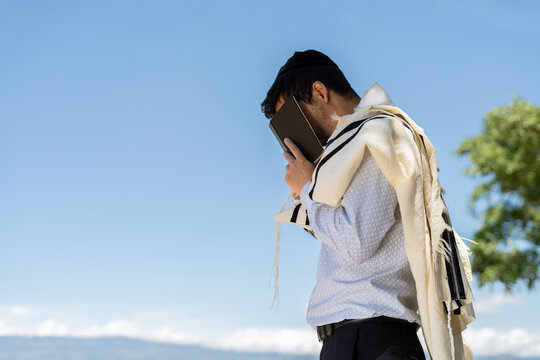 Young Jewish Man Using Tallit and Kippah Covers His Face with Siddur for the Recitation of Shema Israel Outdoors