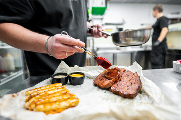 A chef in a professional kitchen glazes a grilled steak with sauce, showcasing culinary artistry. Side dishes are visible in the foreground, capturing the vibrant restaurant atmosphere.