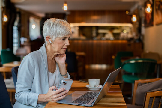 Senior businesswoman working on laptop in a cafe with wireless earbuds