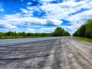 Scenic view of a wide highway bordered by vibrant green trees under a cloudy sky in a rural landscape during daylight