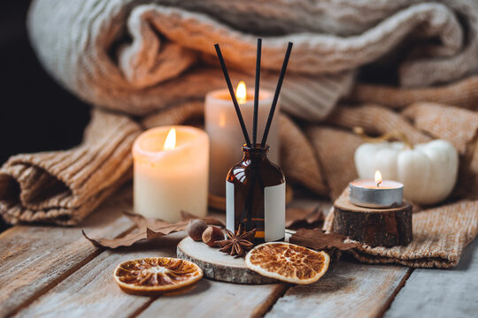 Hello autumn, cozy slow living. Pumpkin, aromatic candle, warm sweaters, dry fall leaves, cinnamon, anise in golden lights with bokeh on white wooden background. Thanksgiving apartment decor