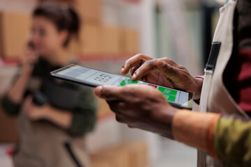 African american worker checking postal form on tablet computer, searching clients parcel before start preparing order. Storehouse employee supervising cardboard boxes in storage room. Close up