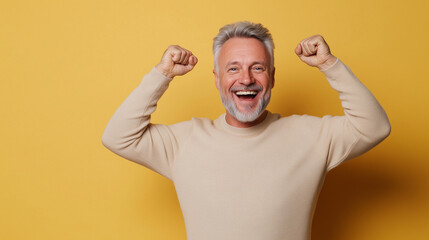 Fototapeta premium A mature man in a light-colored sweater, grinning widely with his hands held high in triumph, isolated against a calm pastel backdrop with copy space around him.