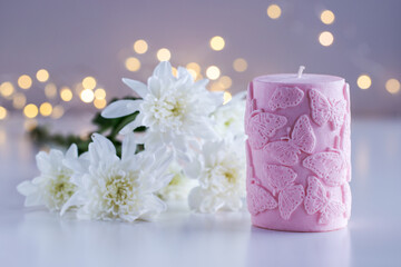 Pink decorative candle with butterflies pattern. Flowers and lights in background with copy space. Home decor. Cozy interior. Soy wax candle making.
