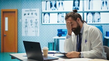 Obraz premium Tired worried doctor working after hours to find the diagnostic for a patient, feeling very stressed and trying to examine x ray results in medical cabinet. Frustrated overworked medic. Camera B.