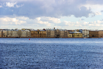 A panoramic view of the historic waterfront of St. Petersburg, Russia, showcasing rows of classic buildings along the Neva River under a partly cloudy sky.