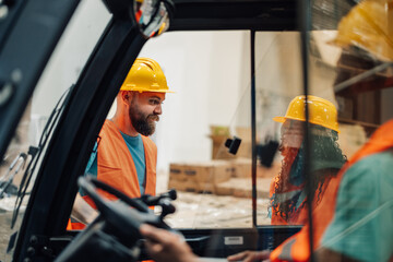 Warehouse workers in hard hats communicating via forklift window.