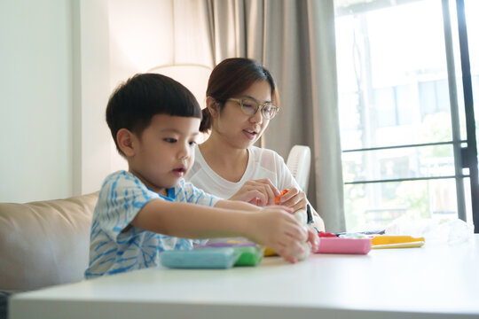 Asian mother and her young son sit at a table, playing with colorful modeling clay together. The boy focuses on his creation while the mother offers guidance in a cozy home setting.