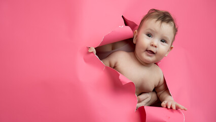 Cute Caucasian newborn baby boy peeks out of a hole in a paper pink background.