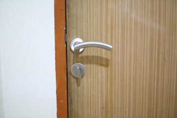 A close-up of a wooden door with a silver handle, slightly ajar, revealing a white wall and an orange frame.