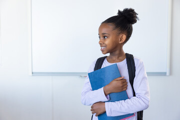 In school, african american girl holding blue notebook and smiling, wearing backpack