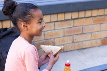 Smiling african american girl eating sandwich and drinking juice during school break outdoors