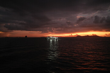 A ferry on a calm sea at sunset with clouds and distant hills in the background