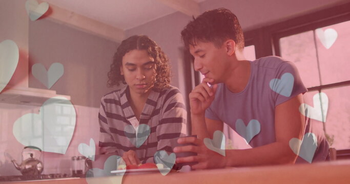 Image of blue hearts over happy diverse gay male couple feeding each other fruit in kitchen