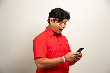 Excited young Asian men celebrate Indonesian independence day on 17 August while holding the Indonesian flag and mobile phone isolated over white background