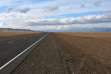 A straight asphalt road goes through the autumn steppe, surrounded by high snow-capped mountains along power line poles.