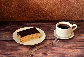 Tiramisu cake and a cup of hot black coffee on a dark wooden table, a fork nearby.