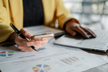 Close-up of a business professional analyzing financial reports with a smartphone and laptop, highlighting modern office work.
