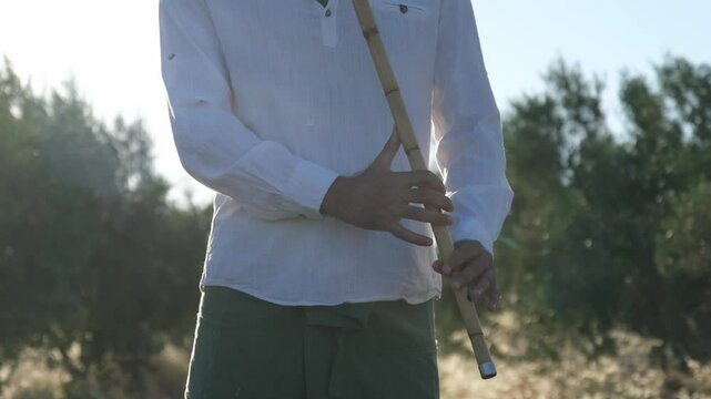 A Middle Eastern man in his 40s plays the ney in an olive grove during sunset, surrounded by nature and tall grasses, creating a peaceful and serene atmosphere