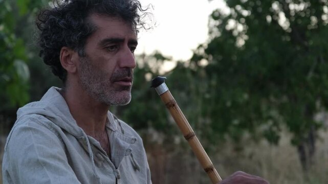 A Middle Eastern man in his 40s plays the ney in an olive grove during sunset, surrounded by nature and tall grasses, creating a peaceful and serene atmosphere