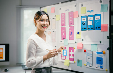Young asian woman happily works as a ux ui designer, smiling while using sticky notes to plan a...