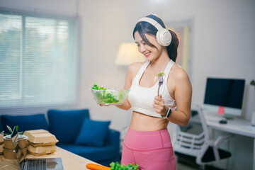 Young woman enjoying a healthy salad after a workout at home, listening to music and smiling in her kitchen. Her fresh meal reflects her commitment to wellness in modern apartment living