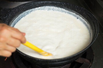 A hand stirring boiled soy milk (soya milk) in black deep pan on  gas stove close up. Homemade creamy soy milk.
